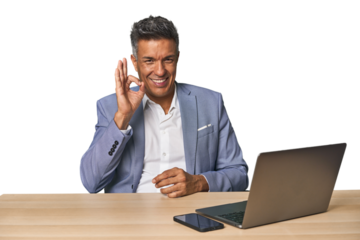 Elegant businessman at desk with laptop winks an eye and holds an okay gesture with hand.
