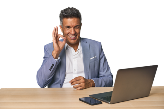Elegant businessman at desk with laptop winks an eye and holds an okay gesture with hand. - Powered by Adobe