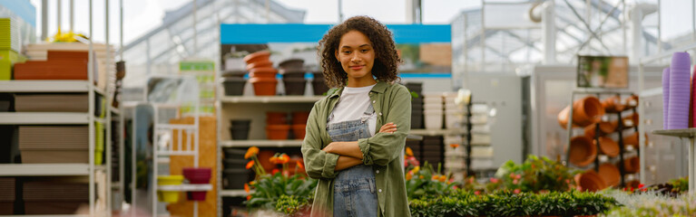 Smiling female garden shop seller standing with crossed hands on flower store background