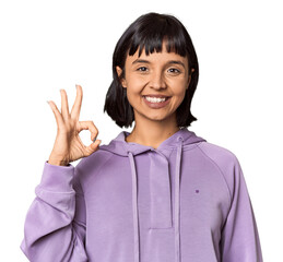 Young Hispanic woman with short black hair in studio winks an eye and holds an okay gesture with hand.