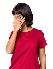 Young Hispanic woman with short black hair in studio blink at the camera through fingers, embarrassed covering face.