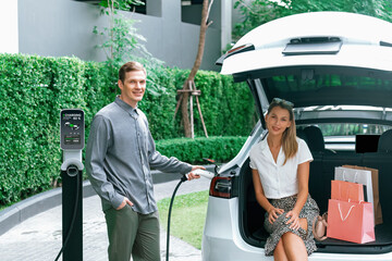 Young couple travel with EV electric car charging in green sustainable city outdoor garden in...