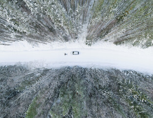 Topshot to snow-covered trees, a snowy road with car, and a person lying in front of it, gazing at the sky