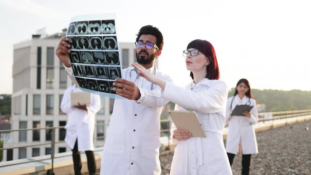 Medical doctor in white lab coat presenting MRI x-ray to young Caucasian nurse while two colleagues relaxing during break outdoors. International group of health care workers at city rooftop.