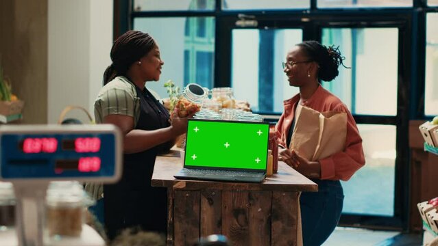 Seller presenting her merchandise next to greenscreen on pc, customer visiting local farmers market to buy bulk products. Merchant recommending grains and pasta, laptop runs isolated chromakey.
