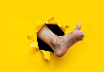 A left woman's foot appears in a torn hole in yellow paper and tries to show a like gesture with...