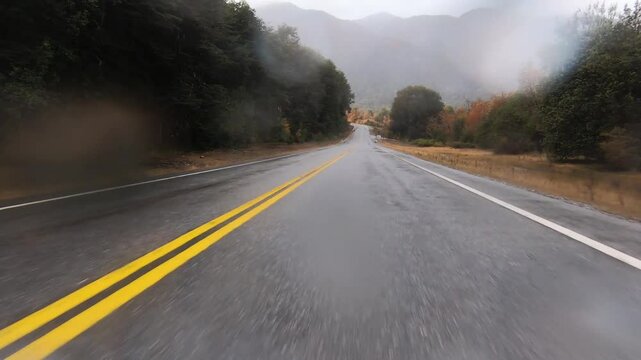 Driving with bad weather. Point of view of a car driving along the asphalt road in the mountain forest. It is raining and the rain drops crush against the lens. 