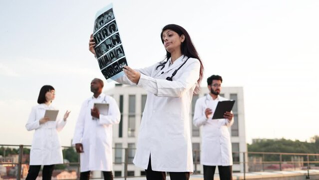 Portrait of ambitious doctor holding x-ray scan of patient during break outdoors hospital. Multiethnic employees of medical clinic working on distant devices with results of examination of patients.