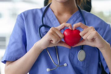 A nurse in blue scrubs holds a red heart in her hands, forming a heart shape around it, symbolizing care. - obrazy, fototapety, plakaty