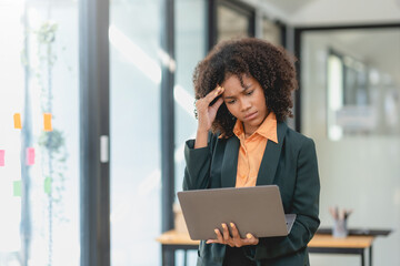 Worried young woman with curly hair looks at a laptop, holding her forehead, in a bright office.