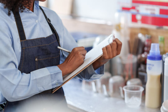 Person in a blue shirt and apron writes on a clipboard in a cafe, surrounded by coffee-making equipment.