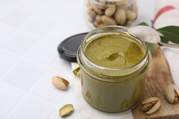 Tasty pistachio cream in jar and nuts on white tiled table, closeup. Space for text