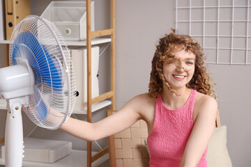 Beautiful happy young woman with electric fan sitting on chair in living room