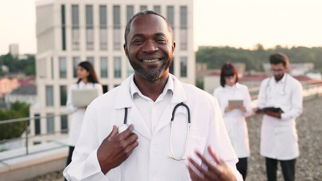Portrait of confident young African doctor in white lab coat posing on camera with stethoscope and smiling. International colleagues of mixed nationality standing behind and operating modern gadgets.