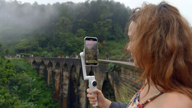 Woman takes on phone with stone bridge and green mountains. Action. Beautiful woman shoots blog in tropical mountains. Woman takes beautiful views of jungle and stone bridge on phone