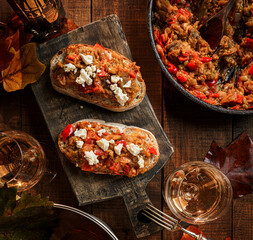 Delicious sandwiches with stewed vegetables and cheese on cutting board, wine in glasses, candles, bottle of wine and autumn leaves on dark wooden background. Wine appetizers, top view