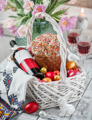 Easter wicker basket with Easter cake, bottle of wine, painted eggs, wine in glasses, flowers tulips on white holiday background with napkin. Easter holiday, close up