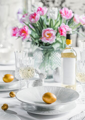 Obraz na płótnie Canvas Easter holidays table, wine in glasses, Easter cake, gold eggs, served plates, flowers tulips, bottle of wine on white background for festive dinner at home. Holiday concept, close up