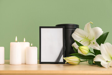 Blank funeral frame, burning candles and beautiful lily flowers on wooden table near color wall