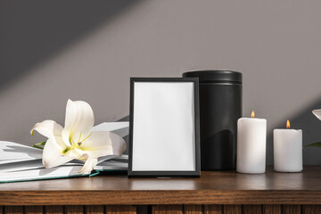 Blank funeral frame, burning candles, mortuary urn and lily flower on wooden cabinet against grey...