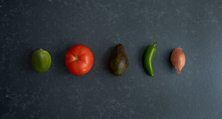 Ingredients for guacamole lined up in a row on a slate gray countertop. There is a lime, a tomato,...