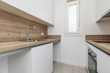 A small kitchen with smooth white furniture without handles and wooden countertops that match the backsplashes, a narrow aluminum window