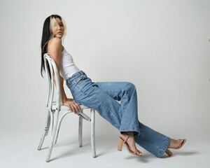 Full length portrait of brunette female asian model wearing casual clothes, white singlet shirt, denim jean pants. Comfortable seated pose on wooden chair. Isolated on white studio background.