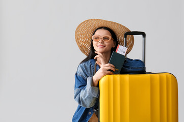 Beautiful young woman in stylish hat with passport, ticket and suitcase on grey background