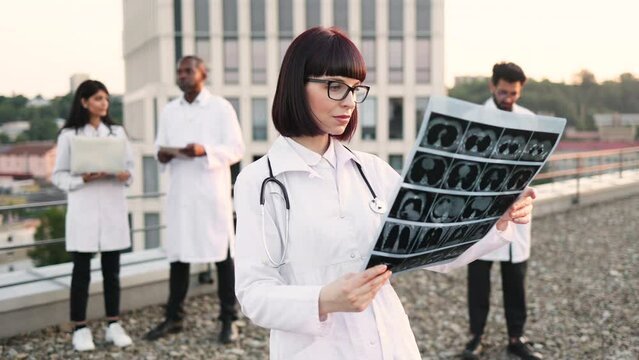 Portrait of Caucasian doctor holding x-ray scan of patient during break outdoors hospital. Multiethnic employees of medical clinic working on distant devices with results of examination of patients.