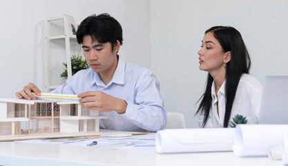 Professional male asian architect using ruler to measure house model length while young beautiful caucasian colleague using laptop to analyzed data on meeting table with house model. Immaculate.