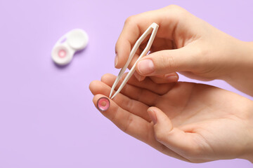 Female hands with tweezers and container with contact lenses on lilac background