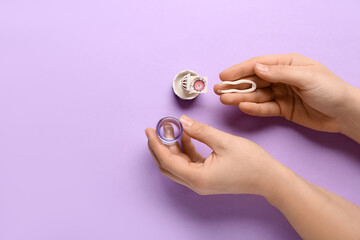 Female hands with cleaning case for contact lenses and tweezers on lilac background