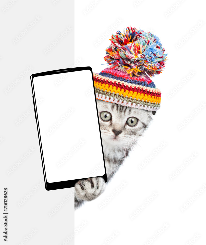 Aufkleber cute kitten wearing warm hat with pompon holds smartphone with white blank screen in it paw above em