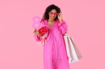 Fashionable African-American woman with bouquet of flowers, figure 8 and shopping bags on pink background. International Women's Day celebration