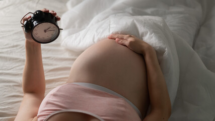 A pregnant woman lies in bed and holds an alarm clock. Close up of the belly.
