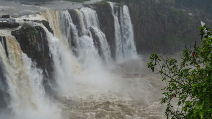 Obraz premium Waterfall flows collapse from a cliff ledge into a stormy river. The water is boiling, foaming, A tourist boat with people is visible in the spray and fog. Iguazu Falls. Brazil