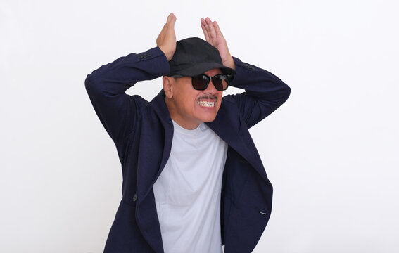 A man wearing a dark blazer over a white t-shirt, hat, and sunglasses was holding his head; angry, mad, disappointed expression