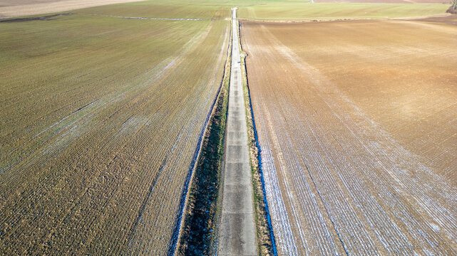 This image offers an aerial view of a long country road stretching into the horizon, dividing two expansive fields. The left field exhibits rows of crops that have been harvested, leaving behind