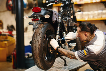 Mechanic in uniform and textile gloves fixing chain of motorcycle - obrazy, fototapety, plakaty