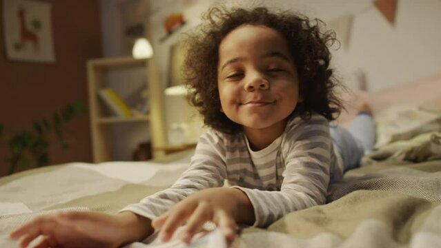 Medium portrait of African American five-year-old curly haired boy happily showing comic book about dinosaurs to camera and smiling while lying on stomach in bed