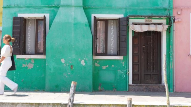 Young female dressed white summer clothes walking by narrow colorful Burano island streets along the water channel. Venice, Veneto region, Italy. 4K footage traveling and vacation concept.