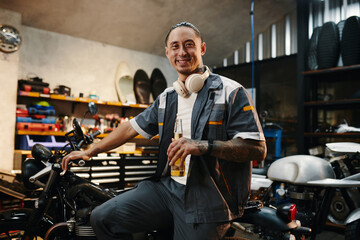 Portrait of happy mechanic with bottle of beer sitting on motorcycle he fixed for client