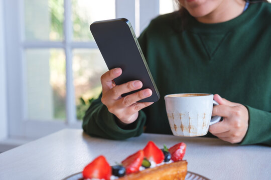 Closeup image of a young woman holding and using mobile phone while drinking coffee in cafe