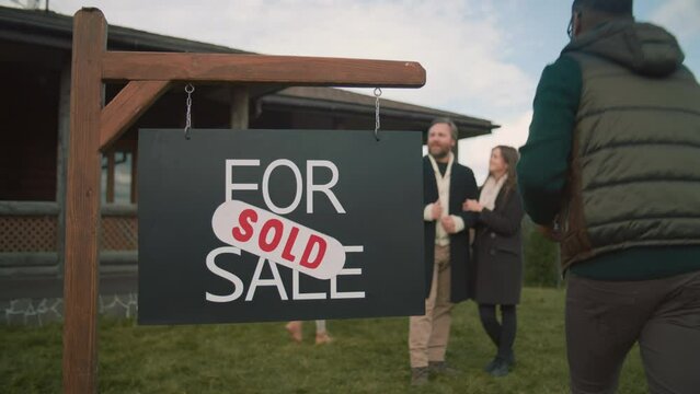 Realtor gives keys of new home to happy family with kids. African American real estate agent comes and puts sold sticker on sign. For sale sign on beautiful green lawn. Residential property on sale.