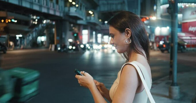On Bangkok's sidewalk woman using phone call taxi waiting amidst auto traffic side view. Taxi call blending with city's rhythm Taxi call quick way to navigate Making evening city travel.