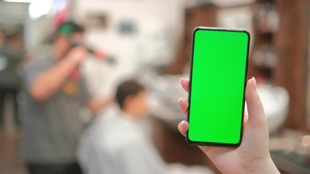 Barbershop. Back view of brunette holding chroma key green screen smartphone watching content. Gadgets and contemporary people concept. 4K