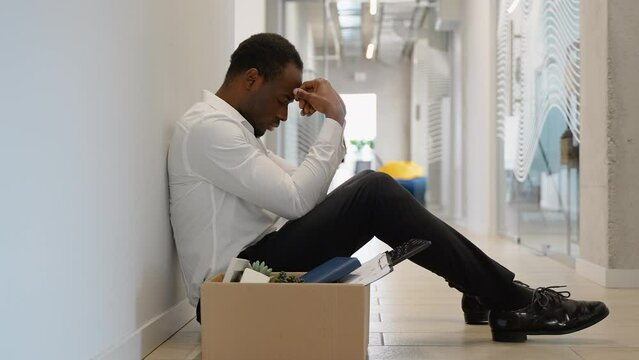 Fired black guy sitting frustrated and upset in the hallway near office with dismissal box