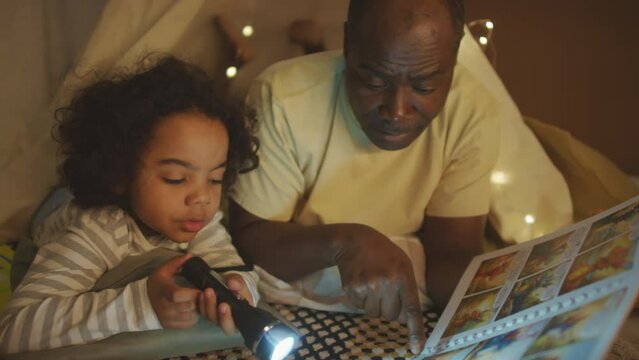 Medium handheld shot of African American dad reading comic strip and pointing at illustrations to kid who holding flashlight and listening, both lying on floor in bedroom at night
