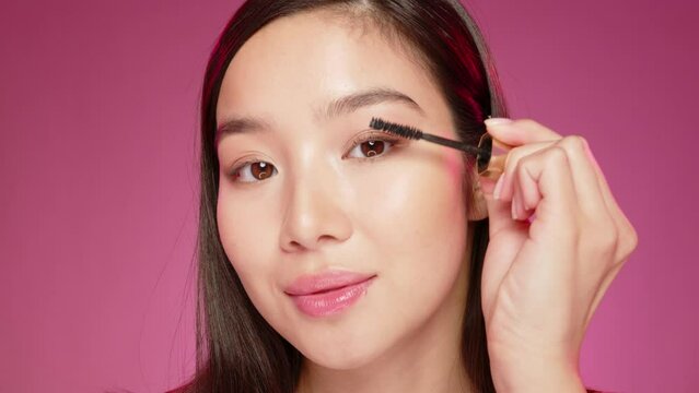 Young Asian girl applying mascara against pink background