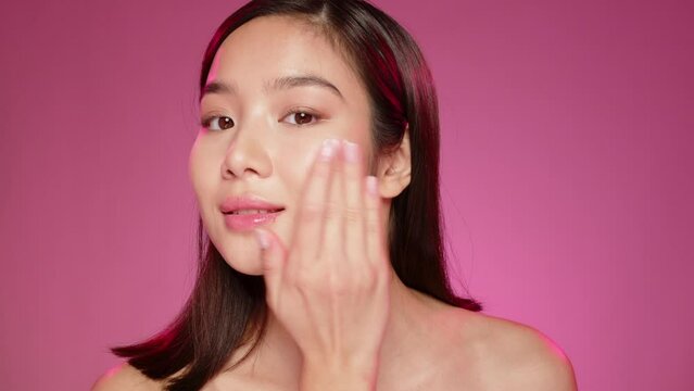 Young Asian girl applying cosmetic cream on face against pink background